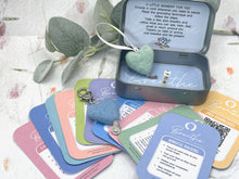 Load image into Gallery viewer, A small open metal tin containing a mint green heart-shaped felt item and a silver keychain, surrounded by colorful printed cards and a second felt piece on a light floral surface.