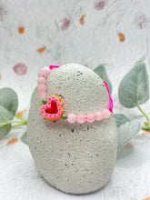 Load image into Gallery viewer, Pink beaded bracelet with a heart charm on a gray stone with leaves in the background
