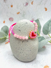 Load image into Gallery viewer, Pink beaded bracelet with a heart charm on a stone surface with leaves in the background
